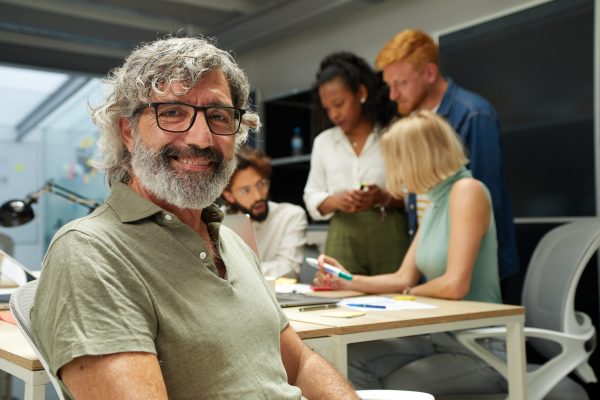 Portrait of an office man smiling and looking at camera. In the background there is a group of employees kipping at work. It is an indoor scene.